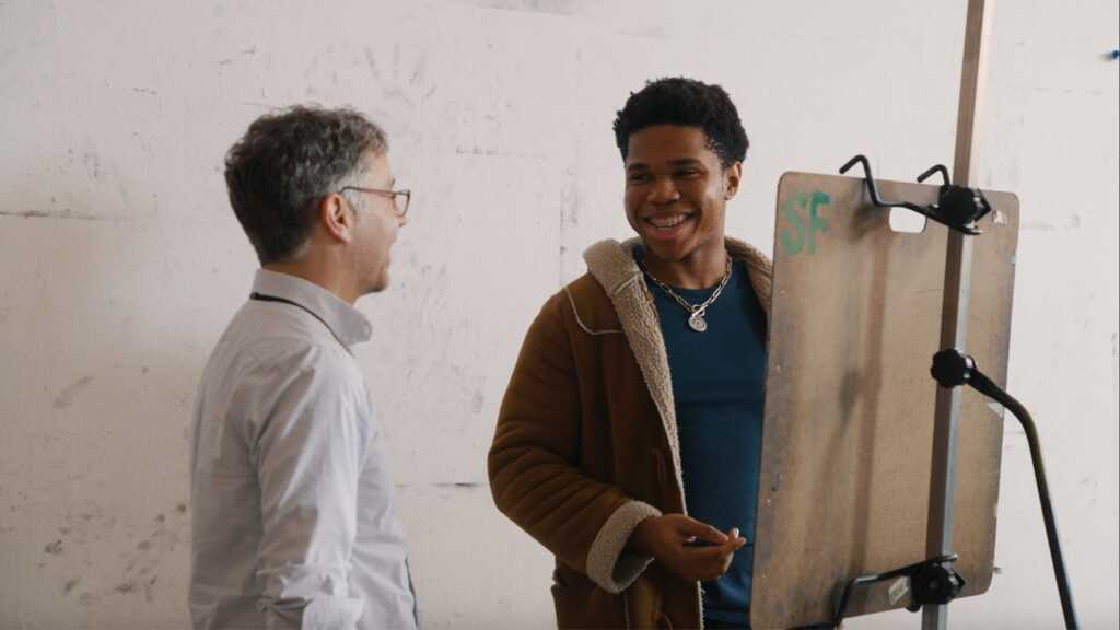 Two people stand and smile at each other. One, a BFA major, holds a pencil by an easel with a sketchpad, suggesting an art lesson or creative discussion in a bright, indoor space.